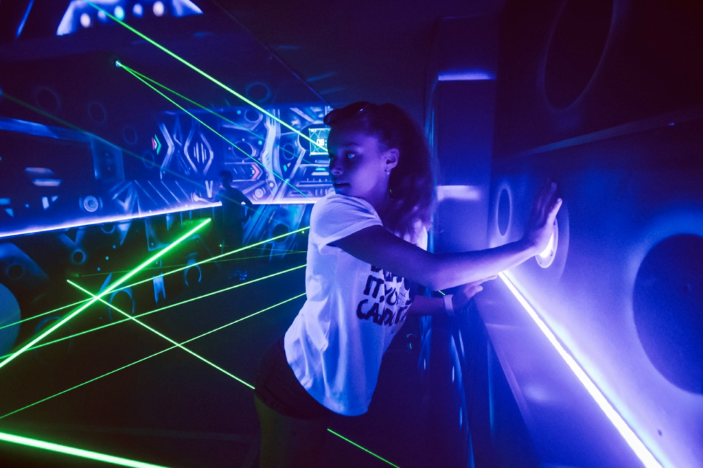 A lady pressing one of the purple illuminated buttons on the wall behind the green lasers in the Laser Labyrinth area