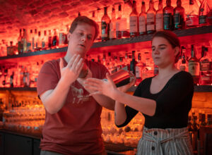 The trainer teaches the lady how to mix drinks in the middle of a bar that doesn't exist